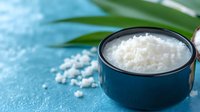 White sea salt crystals in dark blue ceramic bowl on turquoise background with tropical palm leaf, minimalist food styling for healthy cooking and spa.