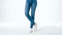Woman wearing blue denim jeans and white sneakers standing against white background, suitable for fashion, retail, and lifestyle marketing materials.