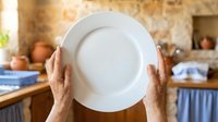 Senior woman with light skin tone holding empty white plate in rustic kitchen with terracotta pottery and wooden countertops, suitable for food service, nutrition, and culinary content.