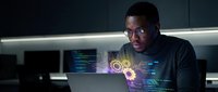 Young African American male programmer working late night coding with holographic data visualization and futuristic interface technology in modern office.