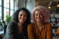 Two cheerful young women with curly hair, one with natural dark curls and one with pink-dyed hair, smiling warmly in casual setting. Friends enjoying time at cafe.