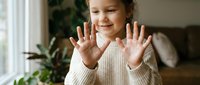 Happy young girl with hands up showing palms in cozy home interior. Child expressing joy and excitement with open gesture in comfortable living space.