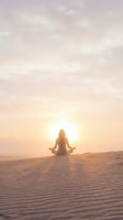 Woman meditating in lotus pose on sand dunes at sunset with warm golden light and peaceful sky for wellness and mindfulness concepts.