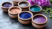 Natural clay bowls with vibrant mineral pigment powders in blue, purple, turquoise, pink and brown shades arranged on dark stone surface with orchid flowers.