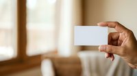Person of African descent holding blank white business card in hand against blurred interior background for branding and corporate identity mockup purposes.