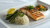 Grilled salmon fillet served with couscous, asparagus, and fresh herbs on white plate. Healthy Mediterranean cuisine with citrus garnish and side vegetables.