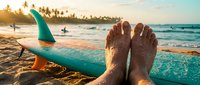 Surfer's feet resting on surfboard at tropical beach during golden hour sunset with ocean waves and palm trees in background, lifestyle and vacation concept.