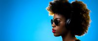 Young African American woman with afro hairstyle wearing sunglasses and headphones in profile against vibrant blue background for music and lifestyle content.