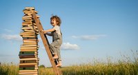 Young woman climbing wooden ladder next to tall stack of books in field under blue sky. Education and knowledge growth concept for learning materials.