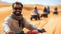 Smiling Middle Eastern man riding quad bike in desert sand dunes, wearing sunglasses and scarf. Adventure tourism group on ATV vehicles in background. Active lifestyle.
