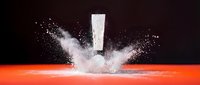 White exclamation mark exploding through orange surface with dramatic powder burst and particles against dark background for impact designs.