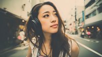 Young Asian woman with headphones listening to music on urban city street at dusk with warm lighting and bokeh background for lifestyle content.