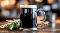 Glass mug of dark craft beer with fresh hop cones and wheat ears on wooden table against blurred pub background, foam head on top of stout ale.