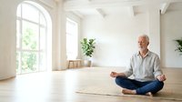 Senior man with light skin tone and bald head, wearing casual button-up shirt and jeans, meditating in lotus position on yoga mat in bright, minimalist studio with large arched windows and potted plants.