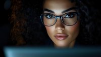 Young woman with dark curly hair and glasses looking at computer screen, professional portrait for business, technology, and education concepts.