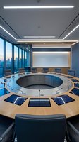 Modern circular conference room with curved wooden table, blue upholstered chairs, linear ceiling lights, floor-to-ceiling windows overlooking city skyline, and central presentation screen for corporate meetings.
