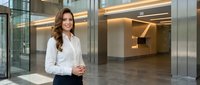 Professional businesswoman in white shirt standing confidently in modern corporate office lobby with glass walls and contemporary architecture.