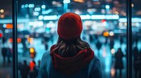Woman in red beanie and sweater looking through window at blurred city lights and crowd during evening, creating atmospheric urban mood with bokeh effect in blue tones.