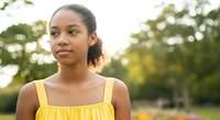 Young woman with medium brown skin tone wearing a yellow sleeveless dress, standing outdoors in a park setting with blurred green foliage background. Portrait suitable for lifestyle, wellness, and diversity concepts.