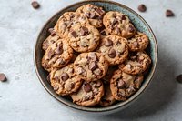Fresh baked chocolate chip cookies piled in rustic ceramic bowl on light marble surface, scattered chocolate chips create appetizing composition.