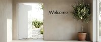 Minimalist interior with open white door leading to sunny garden, welcome text on beige wall, potted olive tree, warm natural light.