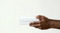 Male hand with dark skin tone holding white boxes or packages against neutral background, suitable for business, delivery, gift, retail, and commerce concepts.