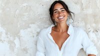 Young Hispanic woman with natural smile wearing white button shirt against textured wall background, expressing joy and confidence in casual lifestyle portrait.