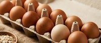 Fresh brown and white chicken eggs in cardboard carton on rustic burlap background with scattered grains, farm-fresh organic produce for cooking and baking.