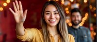 Young Asian woman in yellow sweater waving hello with warm smile against blurred festive background with bokeh lights and male figure in background.