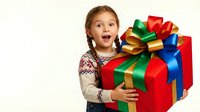 Excited young girl with reddish-brown hair and fair complexion holding large red gift box with colorful ribbons and bows, wearing pink and cream knit sweater, isolated on white background for holiday marketing.