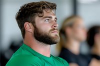 Young Caucasian male with full beard and styled brown hair wearing green t-shirt looking thoughtfully to side, profile view against blurred background in modern setting.
