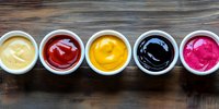Colorful assortment of sauces in white ceramic bowls arranged in row on dark wooden background, including mayonnaise, ketchup, mustard, soy sauce and pink sauce.