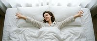 Woman with curly dark hair lying on white bedding with arms stretched out, expressing joy and relaxation. Overhead view for wellness and lifestyle content.