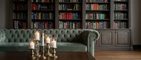 Elegant home library interior with sage green tufted sofa, brass candlesticks with lit candles, and floor-to-ceiling dark wood bookshelves filled with books.