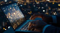 Hands typing on laptop at night with magical glowing screen displaying starry sparkles against blurred city lights background. Creative digital concept.