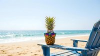 Pineapple wearing sunglasses on beach chair by ocean waves. Tropical summer vacation concept with fruit character relaxing on sandy shore under blue sky.