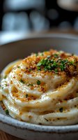 Creamy mashed potatoes swirled in ceramic bowl, topped with crispy breadcrumbs, fresh herbs and black pepper, served on wooden surface with soft background blur.