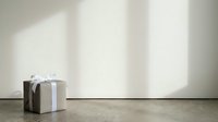 Kraft paper gift box with white ribbon bow on concrete floor against minimalist wall with soft natural light and shadows.
