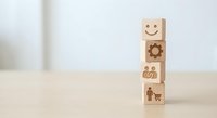 Wooden blocks stacked with business icons including smiley face, gear, dollar sign and human figure on light background for corporate concepts.