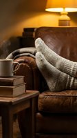Cozy feet in warm knitted socks resting on leather armchair with books and lamp creating comfortable home relaxation atmosphere for evening reading time.