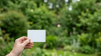 A hand holding a blank white card against a lush green garden background with foliage and natural bokeh, suitable for business cards, marketing mockups, and promotional designs.