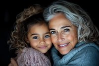 Heartwarming portrait of mature woman with silver hair embracing mixed race granddaughter, both smiling at camera against dark background showing family bond and love.