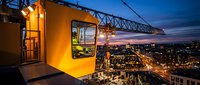 Construction crane operator cabin overlooking illuminated city skyline at dusk with urban lights and evening sky creating dramatic industrial workplace scene.