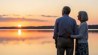 Senior couple embracing while watching golden sunset over calm lake water, romantic evening moment together in peaceful natural setting.