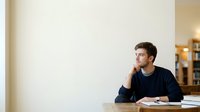 Young adult male, Caucasian ethnicity, wearing casual dark navy sweater, sitting at desk with notebook and pen, hand thoughtfully touching face, looking away contemplatively, suitable for business, education, and lifestyle content.