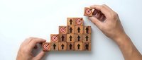 Hands arranging wooden blocks with target icons and human figures in ascending steps, representing team building and goal achievement strategy.