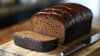 Freshly baked dark chocolate loaf cake with one slice cut, displayed on wooden cutting board with knife.