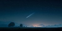 Bright meteor streaking across starry night sky over misty rural landscape with silhouetted trees and distant mountain range at twilight.
