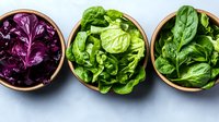 Fresh purple cabbage, butter lettuce, and spinach leaves arranged in wooden bowls on gray background, showcasing variety of leafy greens for healthy eating.