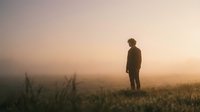 Silhouette of a young man standing alone in a misty field at golden sunrise, fog rolling over grass in warm amber light.